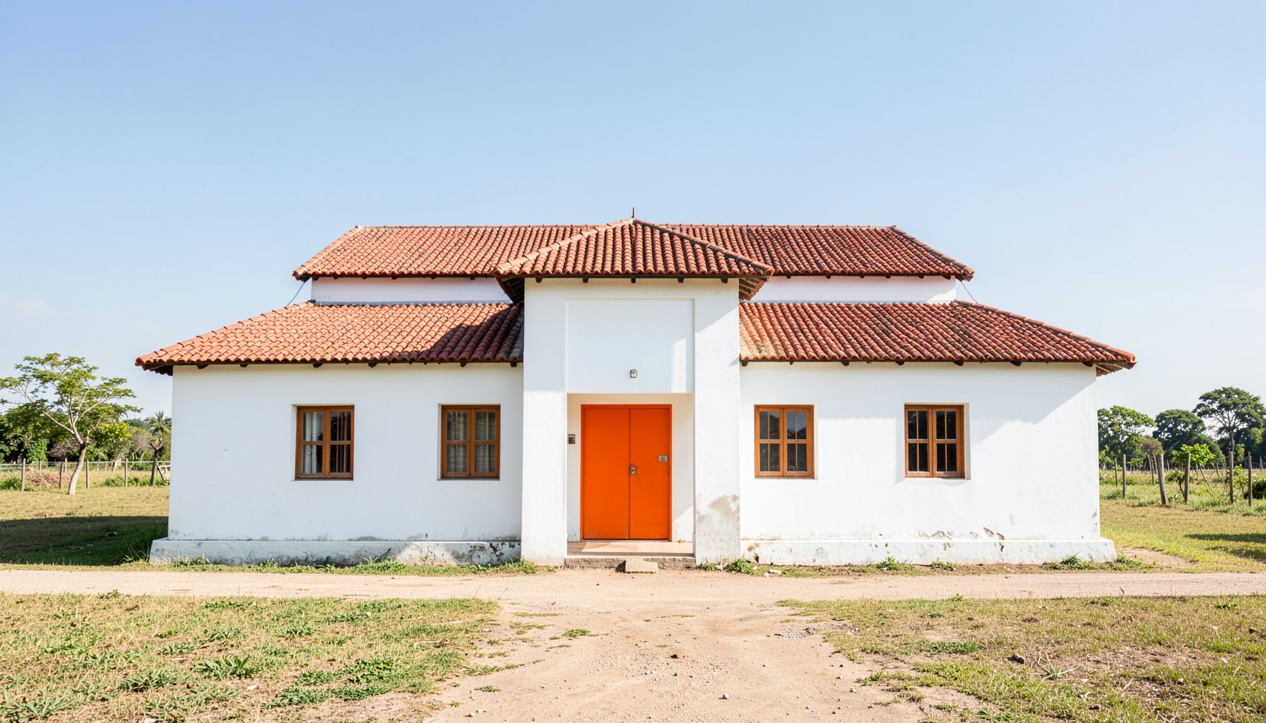 A clean architectural photograph of a modest village meeting hall with strong symmetry, whitewashed walls, and a red tile roof, set against a bright rural landscape under a clear sky.